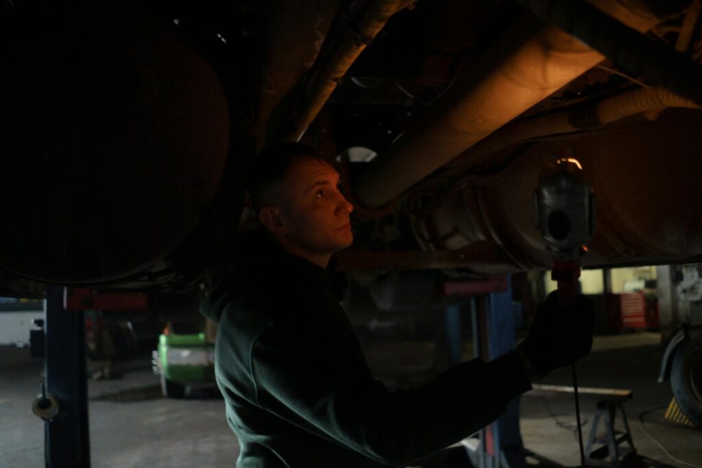 Mechanic working in dimly lit garage, inspecting car's undercarriage with a portable light source.