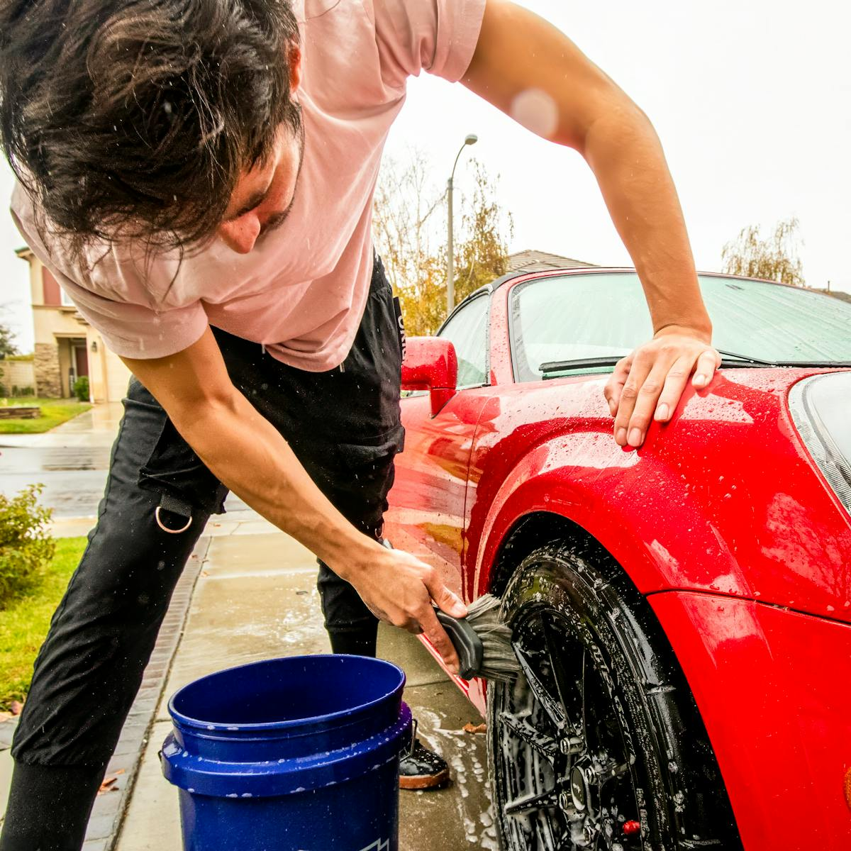 Adult washing a red sports car's wheel using a brush and bucket on a rainy day.