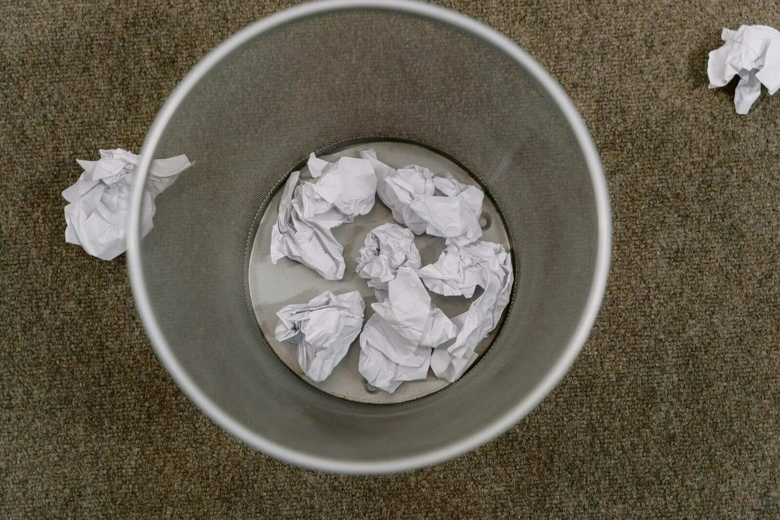 Overhead view of a trash can filled with crumpled paper on a carpeted floor.