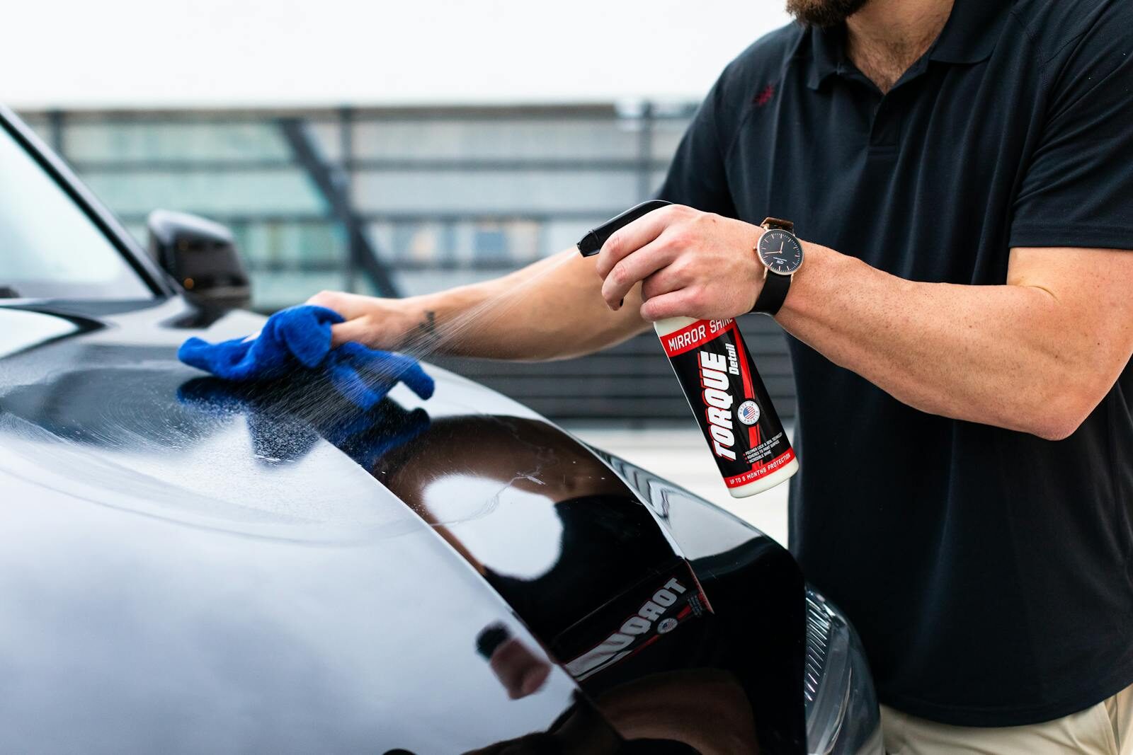 A man cleaning a black car hood with a spray and microfiber cloth.