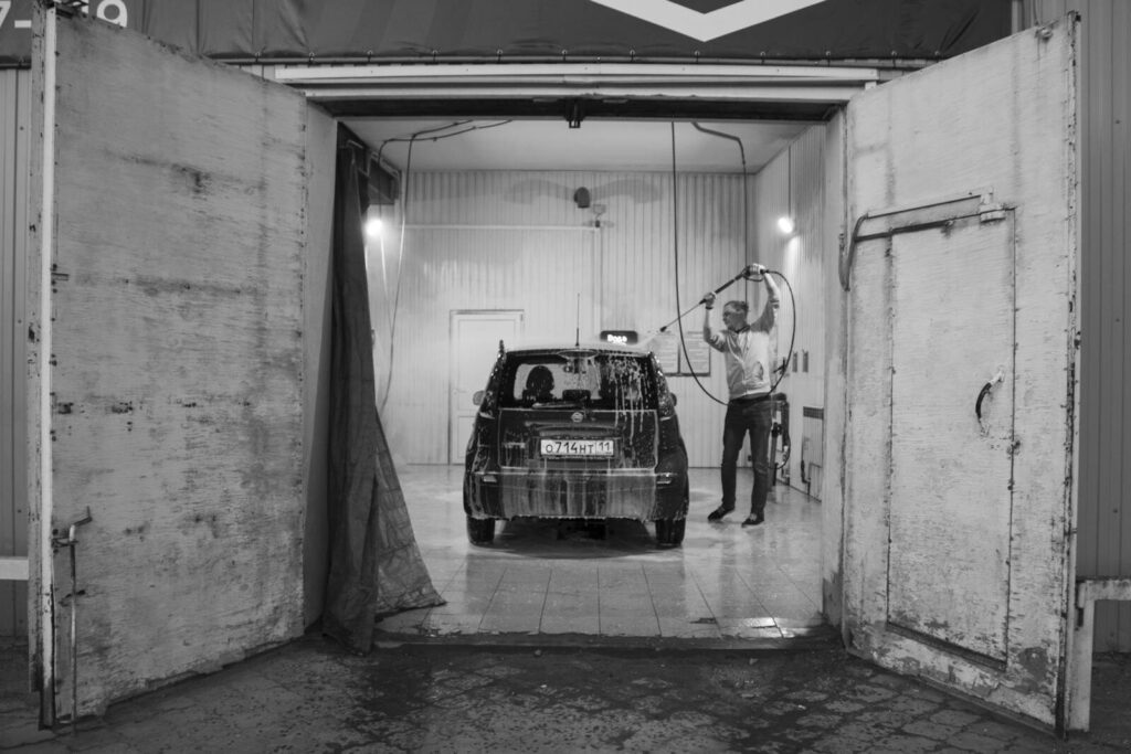 Black and white image of a man washing a car in a garage setting.